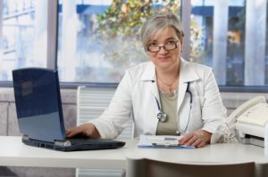 Happy female doctor working at desk in doctors room, using laptop, doing paperwork.